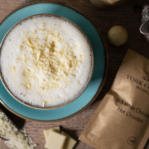 Close up of a white hot chocolate in a blue cup and saucer surrounded by a white chocolate truffle, bar pieces, flakes and a 50g packet.