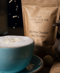 White hot chocolate in a blue cup and saucer with flakes falling and a packet and ornate jug.