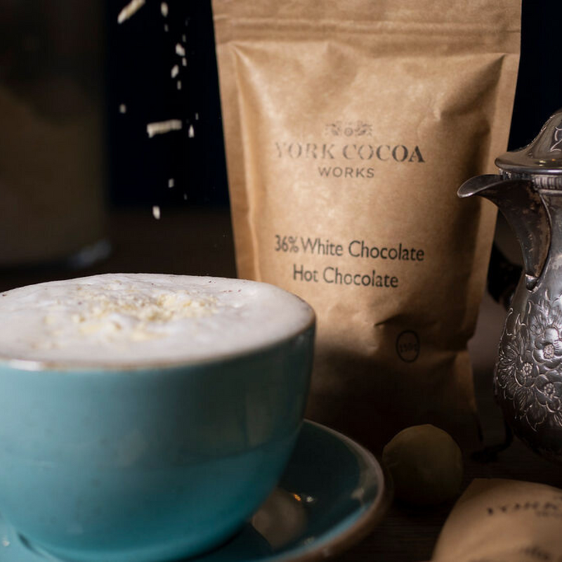 White hot chocolate in a blue cup and saucer with flakes falling and a packet and ornate jug.
