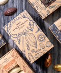 An overhead flat-lay arrangement of multiple small artisanal chocolate boxes on a gray wood surface, with whole and halved chocolate almonds scattered around. The boxes are tan and navy blue with botanical illustrations and visible chocolate windows.