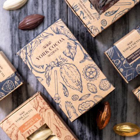 An overhead flat-lay arrangement of multiple small artisanal chocolate boxes on a gray wood surface, with whole and halved chocolate almonds scattered around. The boxes are tan and navy blue with botanical illustrations and visible chocolate windows.