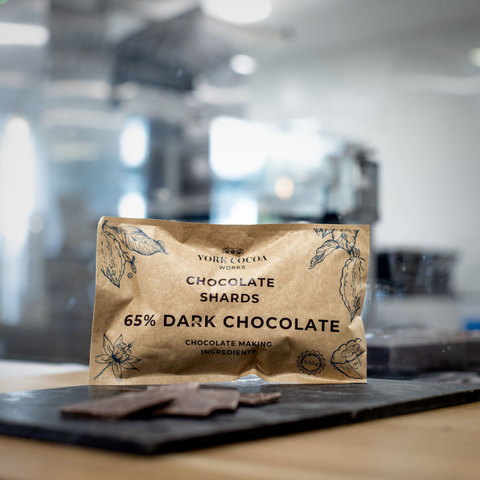 Package of York Cocoa 65% Dark Chocolate Shards on a slate surface and a blurred factory background