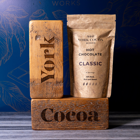 A kraft paper pouch of York Cocoa Works Classic Hot Chocolate stands on a wooden block next to a carved wooden block engraved with 'York' and another block engraved with 'Cocoa', against a dark blue patterned backdrop.