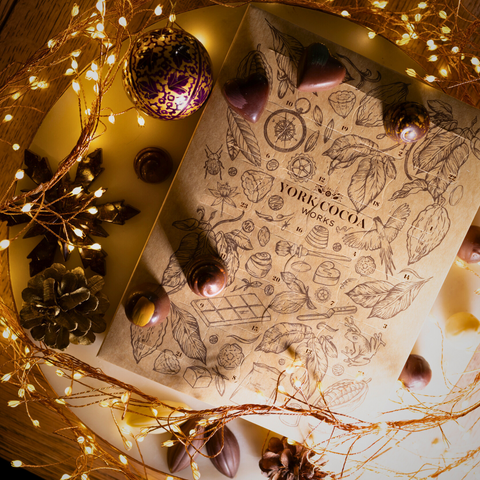Overhead view of Advent Calendar box surrounded by fairylights, festive decorative elements and chocolates on a marble surface.