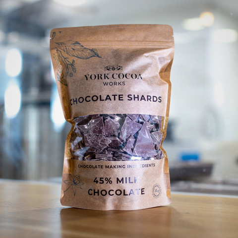 Bag of York Cocoa Works chocolate shards on a wooden surface with a blurred factory background