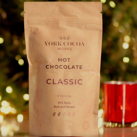 A stand-up pouch of York Cocoa Works Classic Hot Chocolate in front of a blurred background with a Christmas tree, festive lights and a red candle.