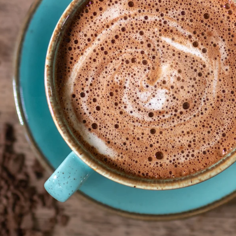 A top-down close-up of a turquoise ceramic mug and saucer filled with a frothy hot chocolate, resting a rustic wooden surface with blurred hot chocolate flakes.