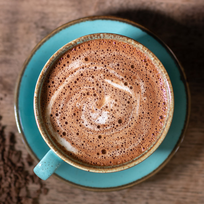 Turquoise mug and saucer with a hot chocolate drink on a wooden surface with loose hot chocolate flakes.