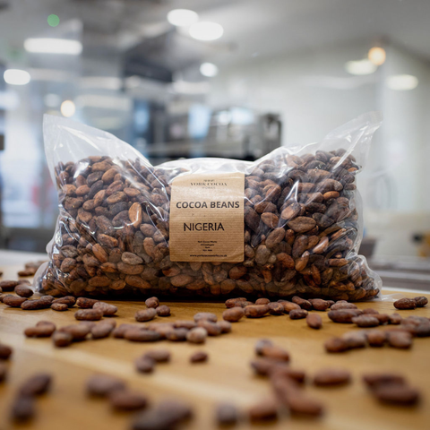 Bag of cocoa beans with a label indicating 'Nigeria' on a wooden surface in front of a blurred factory background.