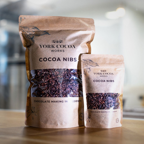 Two packages of York Cocoa Works cocoa nibs on a wooden surface with a blurred factory background.