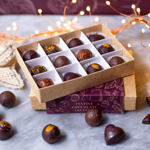 An open box of assorted chocolate truffles in a kraft paper tray, with several chocolate truffles scattered around on a light marble surface. The box lid is placed on top of a branded box that reads 'Festive Chocolate Truffles.