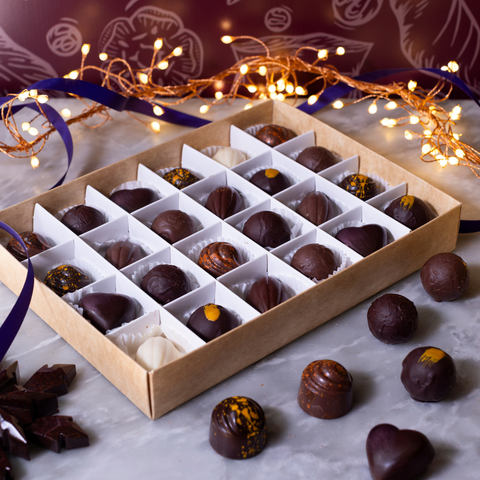 An open kraft gift box filled with assorted chocolate truffles and a few loose chocolates arranged on a marble surface, with warm fairy lights and purple ribbon in the background.