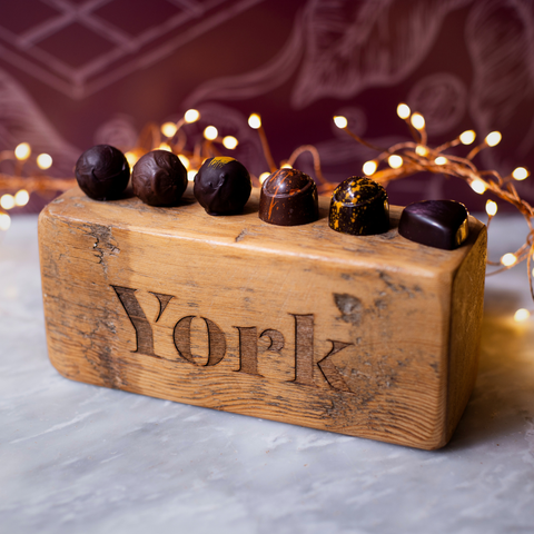 A wooden block engraved with the word 'York' holds six chocolate truffles, photographed on a light marble surface with warm fairy lights and a patterned plum backdrop.