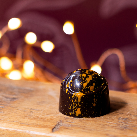 Close up of a mince pie chocolate truffle on a wooden surface surrounded by fairylights and a blurred plum background.