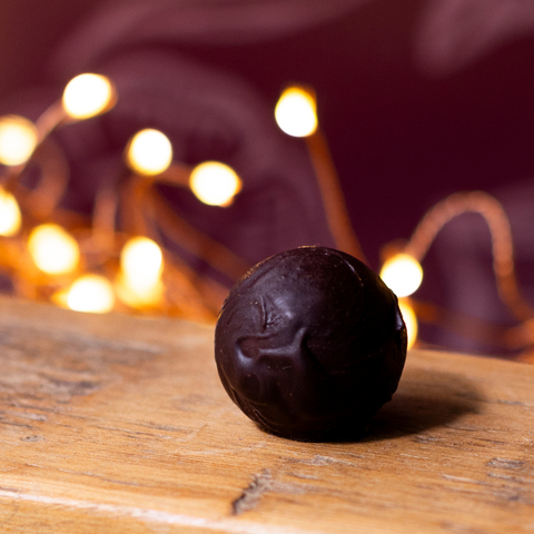 A single dark chocolate truffle sphere resting on a wooden surface with warm fairylights blurred in the background.