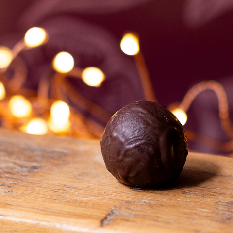A single milk chocolate truffle sphere resting on a wooden surface with warm fairy lights blurred in the background.