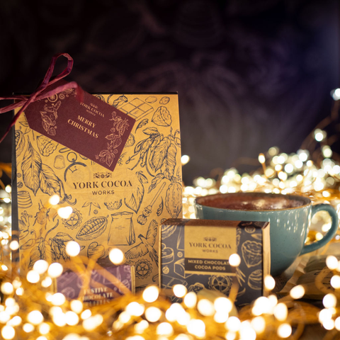 A gift set of York Cocoa Works Christmas treats, featuring a decorated gift box with a plum 'Merry Christmas' tag, a smaller box of mixed cocoa pods and festive chocoalte truffles and a blue mug filled with hot chocolate, surrounded by warm fairy lights and a dark blue background.