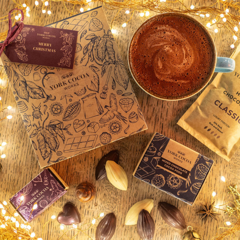 A styled overhead arrangement including a kraft gift box with a "Merry Christmas" tag, a cup of hot chocolate, a hot chocolate packet, a box of mixed cocoa pods and festive truffles with loose chocolates, festive decorations and fairylights on a wooden surface.
