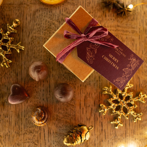 Kraft gift box with a plum ribbon and 'Merry Christmas' tag on a wooden surface with four chocolates and festive decorative elements. 
