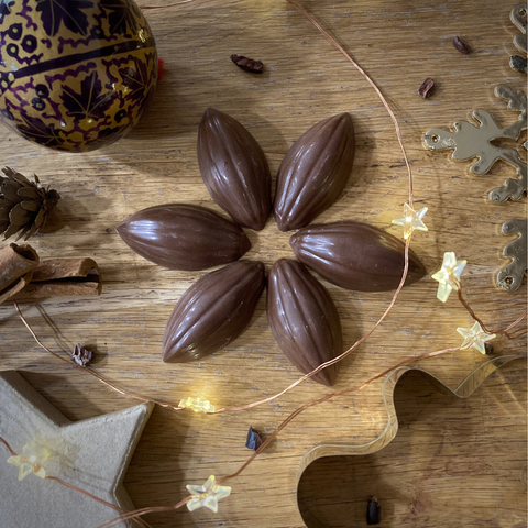 Six Gingerbread Chocolate Cocoa Pods on a wooden surface, surrounded by cocoa nibs and festive decorations.
